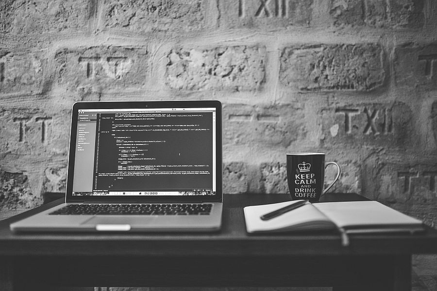 Black and white picture showing a desk with a laptop and a coffee cup in front of a masonry wall